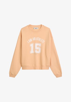 Bluzka z długim rękawem w kolorze brzoskwiniowym, ze ściągaczami u rękawów i okrągłym dekoltem. Na przodzie znajduje się biała grafika z napisem: "JESTEM SPORTOWCEM" oraz wyraźnie widoczną cyfrą "15".