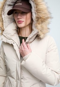 Giacca invernale beige puffa con cappuccio foderato in pelliccia, dotata di zip e chiusure a scatto. Abbinata a un cappello marrone con testo ricamato.