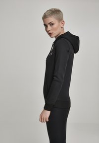 Black hooded sweatshirt with a front pocket, ribbed cuffs, and hem. Smooth texture and relaxed fit, displayed on a model from the side.