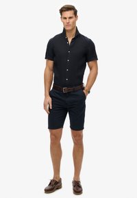 Granatowa koszula z krótkim rękawem zapinana na guziki, zestawiona z ciemnymi szortami i brązowymi skórzanymi butami żeglarskimi. Brązowy pasek i zegarek jako dodatki.