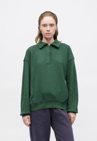 Suéter verde texturizado con cuello y media cremallera. Presenta laterales acanalados y un ajuste relajado, combinado con pantalones morados holgados.