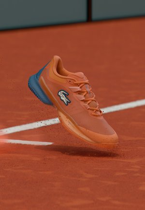 Narančasti sportski tenis sa logom marke u zraku iznad teniskog terena od zemlje, podižući crvene čestice zemlje blizu bijele linije terena.