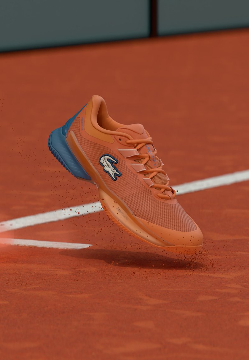 Πορτοκαλί αθλητικό παπούτσι με λογότυπο μάρκας στον αέρα πάνω από γήπεδο τένις με χωμάτινη επιφάνεια, σηκώνοντας κόκκινα σωματίδια χώματος κοντά στη λευκή γραμμή του γηπέδου.