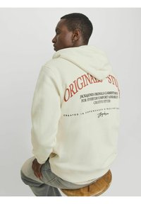 Krämfärgad oversized hoodie med dragsko, med röd text på baksidan. Slätt tyg med ribbade cuffs, bärs över ljusblå jeans.