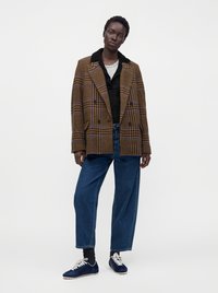 Personne debout portant un blazer marron à carreaux, une chemise noire, un sous-vêtement blanc, un jean bleu, des chaussettes noires et des baskets bleues sur un fond uni.