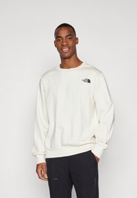 Creme sweatshirt med rund hals og ribbede manchetter, med et sort logo på venstre bryst. Båret med mørke bukser.
