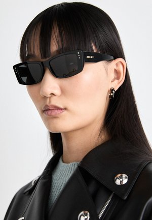 Óculos de sol retangulares pretos com lentes escuras, apresentando pequenos pontos de destaque na armação. Usados com uma jaqueta de couro preta e uma blusa de clara.