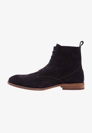 Bottines pour hommes en suède noir avec un bout arrondi, design simple à lacets et semelle marron contrastante. Détails de couture subtils sur les côtés.