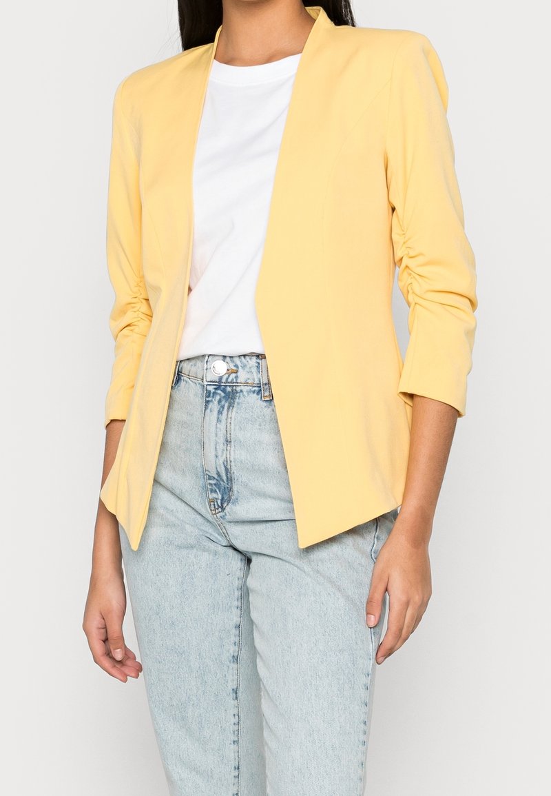 Veste ajustée jaune à manches trois-quarts, avec une texture lisse et une coupe moderne, portée sur un t-shirt blanc et un jean en denim clair.