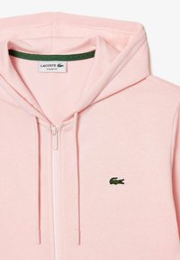 Světlá růžová mikina na zip s kapucí a šňůrkami, zelené vyšití loga Lacoste na levé straně hrudi a štítek uvnitř límce s nápisem "Classic Fit".