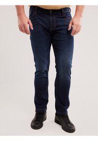 Bărbat purtând blugi slim-fit de culoare albastru închis și adidași negri, stând în picioare pe un fundal deschis simplu.