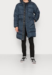 Marinblå puffjacka med huva, vertikal quiltad design, fickor med dragkedja och en svart drajdkedja framtill. Buren över svarta byxor och sneakers.