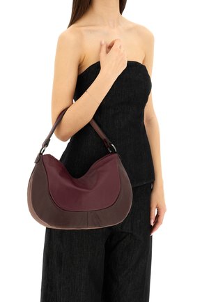 Borsa hobo realizzata in pelle borgogna testurizzata con un design curvo e una tracolla marrone scuro, portata sotto il braccio contro un outfit nero.