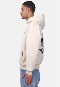 Beige Kapuzenpulli mit einem großen schwarzen Grafikdesign am linken Ärmel. Verfügt über eine Vordertasche, eine Kordelzugkapuze und gerippte Bündchen.