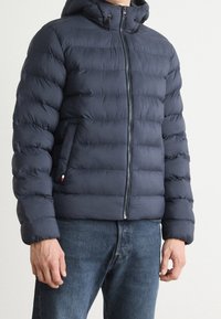 Navy blue puffer jacket with a zippered front, hood, and textured quilted pattern. Features side pockets and a small brand logo accent.