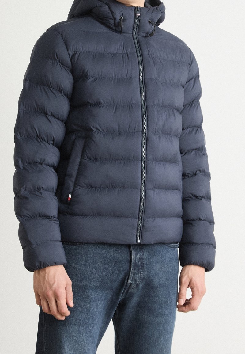 Navy blue puffer jacket with a zippered front, hood, and textured quilted pattern. Features side pockets and a small brand logo accent.