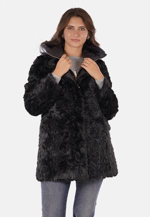 Cappotto in pelliccia sintetica nera con chiusura a zip, colletto ampio e una finitura morbida e testurizzata. Indossato sopra un top grigio e abbinato a un denim.