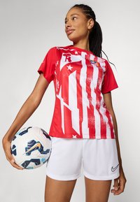 Rød fotballtrøye med grafisk design av hvite striper og stjerner, sammen med hvite shorts med Nike-logo; holder en mønstrete fotball.