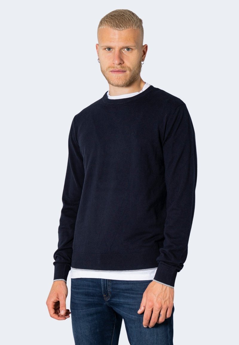 Maglione a manica lunga blu navy realizzato in tessuto morbido, con colletto rotondo, polsini a coste e una texture a maglia fine. Indossato sopra una camicia bianca.