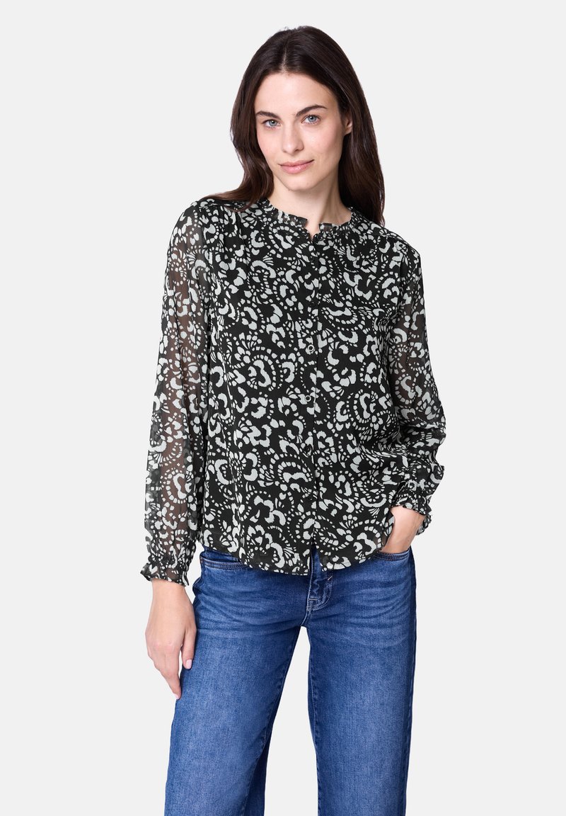 Schwarz-weiß gemusterte Bluse mit langen Ärmeln, hohem Kragen und Knopfleiste. Aus leichtem, transparentem Stoff. Kombiniert mit blauen Jeans.