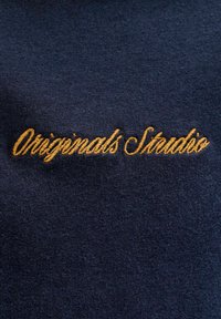 Ύφασμα σε ναυτικό μπλε με καλλιγραφική χρυσή κεντητή επιγραφή που γράφει "Originals Studio" στο κέντρο της υφασμάτινης επιφάνειας.