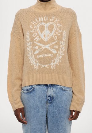 Beige pooloneule, jossa valkoinen rauhan sydän, neulepuikot, laakeriseppeleen kuvio ja teksti "MOECHINO JEANS 1986 2023 KÄSINKUDOTTU", yhdistetty sinisiin farkkuihin.