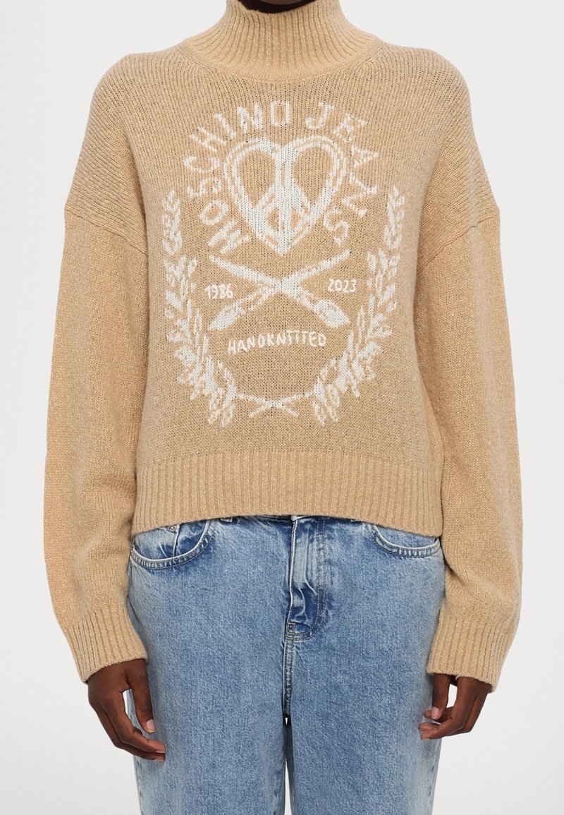 Pull à col roulé beige avec un cœur blanc de la paix, des aiguilles à tricoter, un motif de laurier et le texte "MOECHINO JEANS 1986 2023 FAIT MAIN", porté avec un jean bleu.