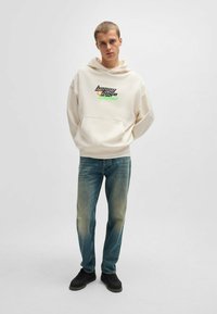 Felpa crema con tasca frontale e testo grafico, abbinata a jeans blu sbiaditi e sneaker nere, in piedi contro uno sfondo neutro.