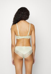 Mulher com cabelo longo e encaracolado, vestindo um soutien verde claro com renda e uma combinação de panties, de costas para a câmara num fundo branco.