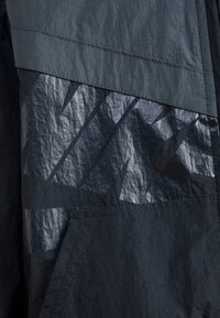 Tmavá, strukturovaná bunda s lesklým černým grafickým vzorem, prošívanými detaily a kombinací matných a lesklých ploch.