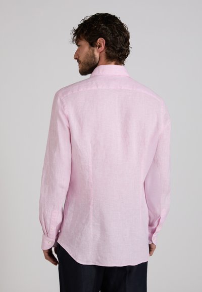 Uomo con capelli ricci scuri che indossa una camicia a maniche lunghe rosa chiaro, visto da dietro su uno sfondo grigio uniforme.