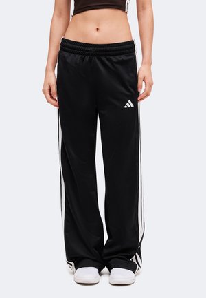 Personne portant un pantalon de survêtement Adidas noir avec des bandes blanches et un logo, associé à des chaussures blanches, montré de la taille aux pieds.