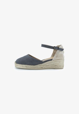 Espadrilles de lona gris con una suela de cuña de yute y una parte trasera de tela texturizada. Cuenta con una correa en el tobillo con un hebilla de metal.