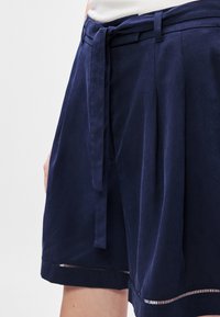 Shorts bleu marine en tissu doux, dotés d'une ceinture, de plis et de détails découpés décoratifs le long de l'ourlet inférieur.