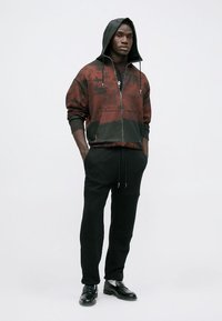 Giacca bomber con cappuccio in gradiente rosso e nero, con chiusura a zip e tasche frontali, abbinata a jogger neri e mocassini neri.