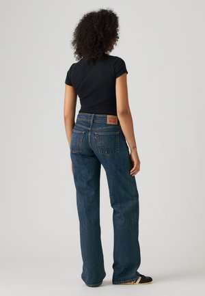 Femme aux cheveux bouclés portant un haut noir à manches courtes et un jean bleu taille haute, debout de dos sur un fond uni.
