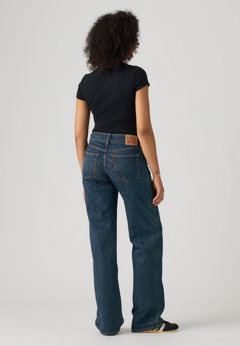 Femme aux cheveux bouclés portant un haut noir à manches courtes et un jean bleu taille haute, debout de dos sur un fond uni.