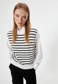 Suéter sin mangas de punto a rayas blancas y negras sobre una camisa blanca con cuello, con un dobladillo acanalado y una textura suave.