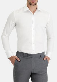 Camisa blanca de manga larga con botones en el delantero, cuello clásico y tejido suave, combinada con pantalones de vestir grises con frente plano y bolsillos.