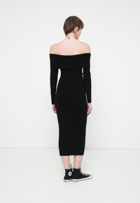 Rochie neagră cu umeri descoperiți, mâneci lungi, siluetă ajustată și lungime până la jumătatea gambei. Asortată cu adidași negri cu talpă înaltă.