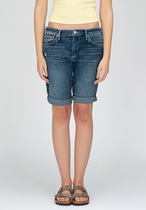 Person trägt hochgekrempelte dunkelblaue Jeansshorts, gelbes bauchfreies Top und braune Sandalen und steht auf einem einfarbigen Hintergrund frontal.
