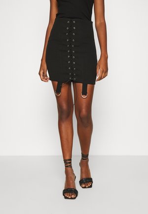Femme portant une mini-jupe noire à lacets avec anneaux argentés, des talons noirs à brides et un haut noir sans manches, debout sur un fond blanc.