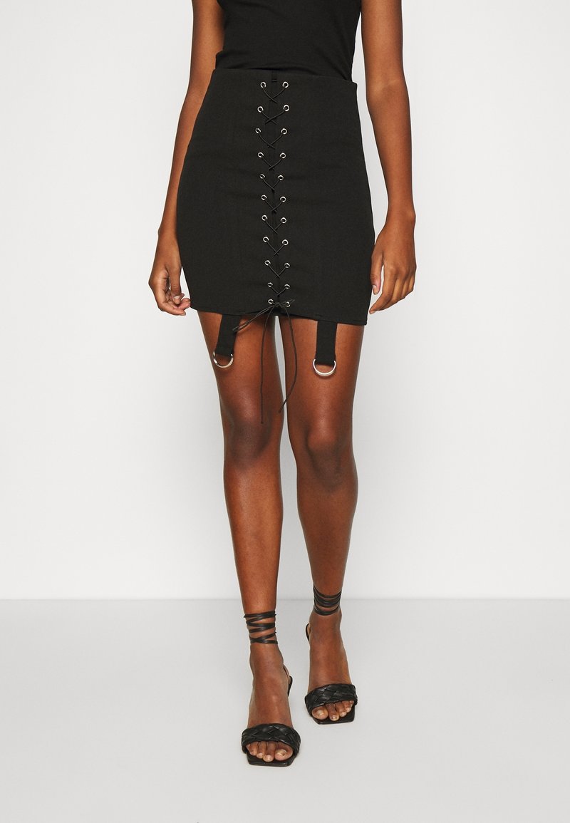 Femme portant une mini-jupe noire à lacets avec anneaux argentés, des talons noirs à brides et un haut noir sans manches, debout sur un fond blanc.