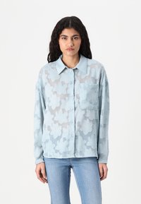 Blusa azul clara transparente com um padrão floral, frente abotoada e um único bolso no peito. Mangas longas com bainhas ligeiramente ajustadas.