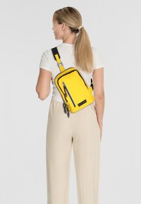 Bolso cruzado amarillo de nailon con un bolsillo con cremallera, ribete negro y correa ajustable, llevado en la espalda, combinado con pantalones de color claro.