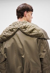 Młody mężczyzna widziany od tyłu, ubrany w zieloną kurtkę z pikowanym, odpinanym kołnierzem, częściowo rozpiętą i spoczywającą na ramionach.