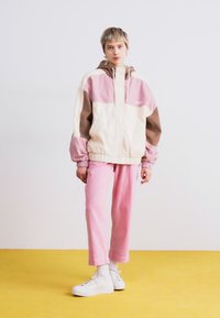 Farvet jakke i creme, pink og brun; elastiske manchetter; parret med pink bukser og hvide højhælede sneakers på en gul gulv.