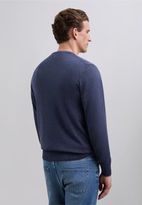Mand med kort brunt hår iført en blå langærmet sweater og blå jeans, vendende til højre foran en ensfarvet lys baggrund.