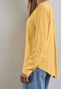 Gul långärmad blus med en slät textur, med rundad nederkant, knäppta manschetter och en avslappnad passform, som bärs tillsammans med blå jeans.
