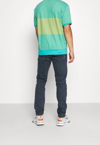 Camisa de manga corta de color turquesa y amarillo con un diseño estampado, con un dobladillo sólido en turquesa, combinada con jeans oscuros y zapatillas blancas con detalles en naranja.